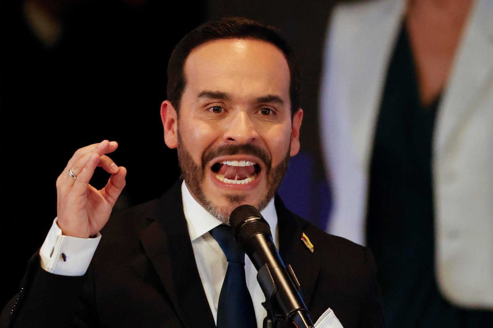 Ao centro, o presidente colombiano Abelardo de la Espirella. Ele é um homem branco, de cabelo curto e preto. Usa um terno preto,está de boca aberta, e olhando para a frente, atrás de um microfone.
