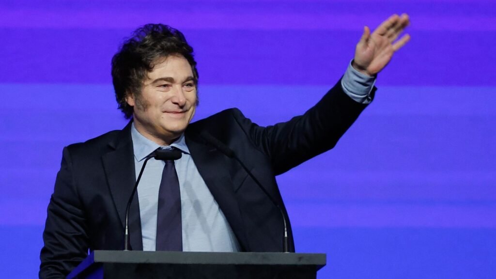 Milei assina decreto contra ódio após ofender Brasil Ao centro, o presidente Javier Milei de terno e gravata pretos e camisa social azul marinho. Ele é um homem branco, sorrindo e com a mão esquerda erguida. Está atrás de uma tribuna com microfone.