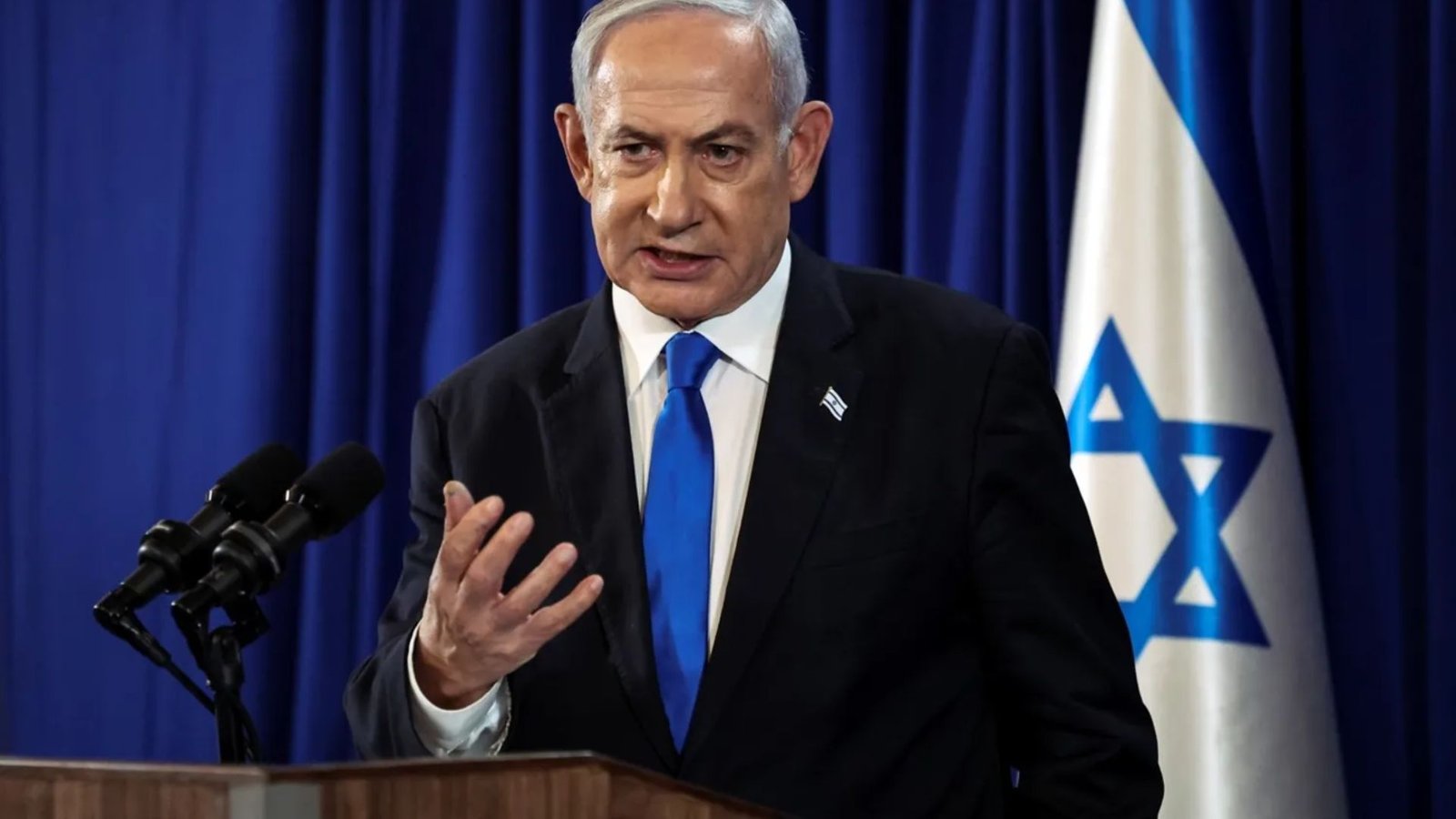 Ao centro, o primeiro-ministro de Israel Benjamin Netanyahu. Ele é um homem de pele e cabelo brancos, usa um terno preto, com gravata azul escuro e camisa social branca. À sua frente, um pulpito com microfones. Ao lado direito, a bandeira de Israel.