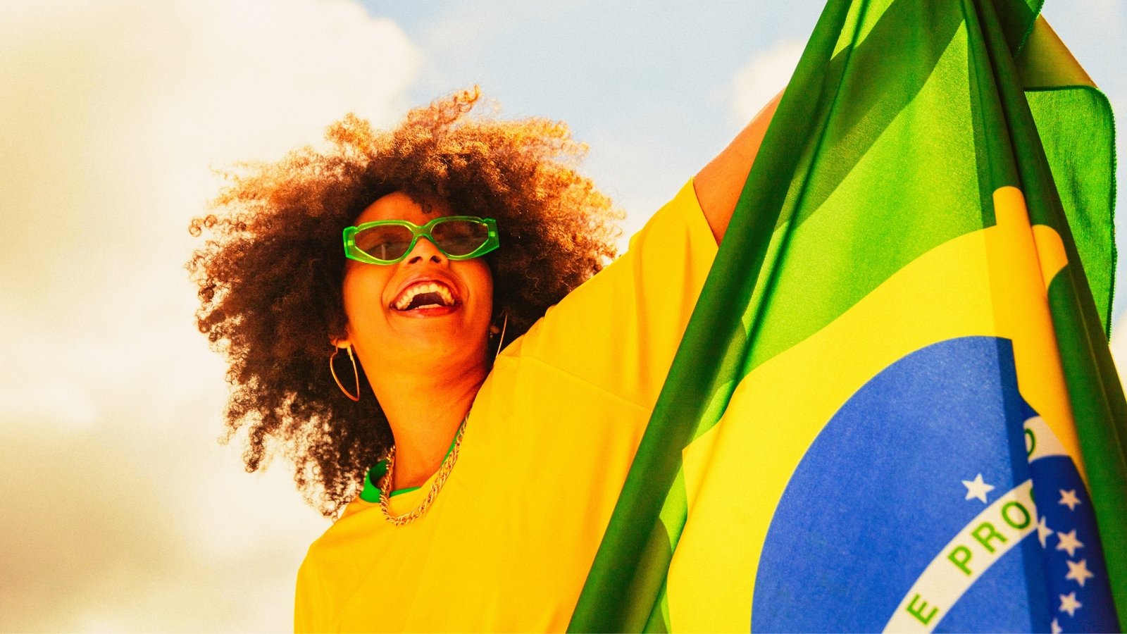 Ao centro, uma torcedora da seleção brasileira, negra e de cabelos crespos sorri e olha para cima. Ela usa óculos escuros com detalhes verdes. Segura uma bandeira do brasil do centro para a direita.